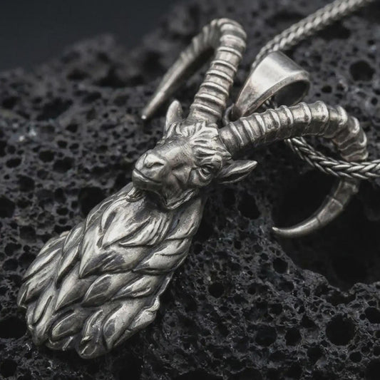 Oxidized ram horn pendant necklace on textured volcanic rock, close-up studio shot highlighting the sculpted goat head, curved horns, leaf-like beard detail, and antique silver contrast.