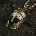 Load image into Gallery viewer, Bronze Spartan helmet pendant necklace on textured volcanic rock, close-up studio shot highlighting the cracked warrior helmet, open face guard, battle-worn texture, and antique silver contrast.
