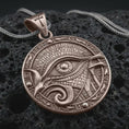 Load image into Gallery viewer, A Bronze Eye of Horus pendant rests on dark volcanic rock in a macro studio close-up, showing Egyptian-inspired details and a bold protection symbol.
