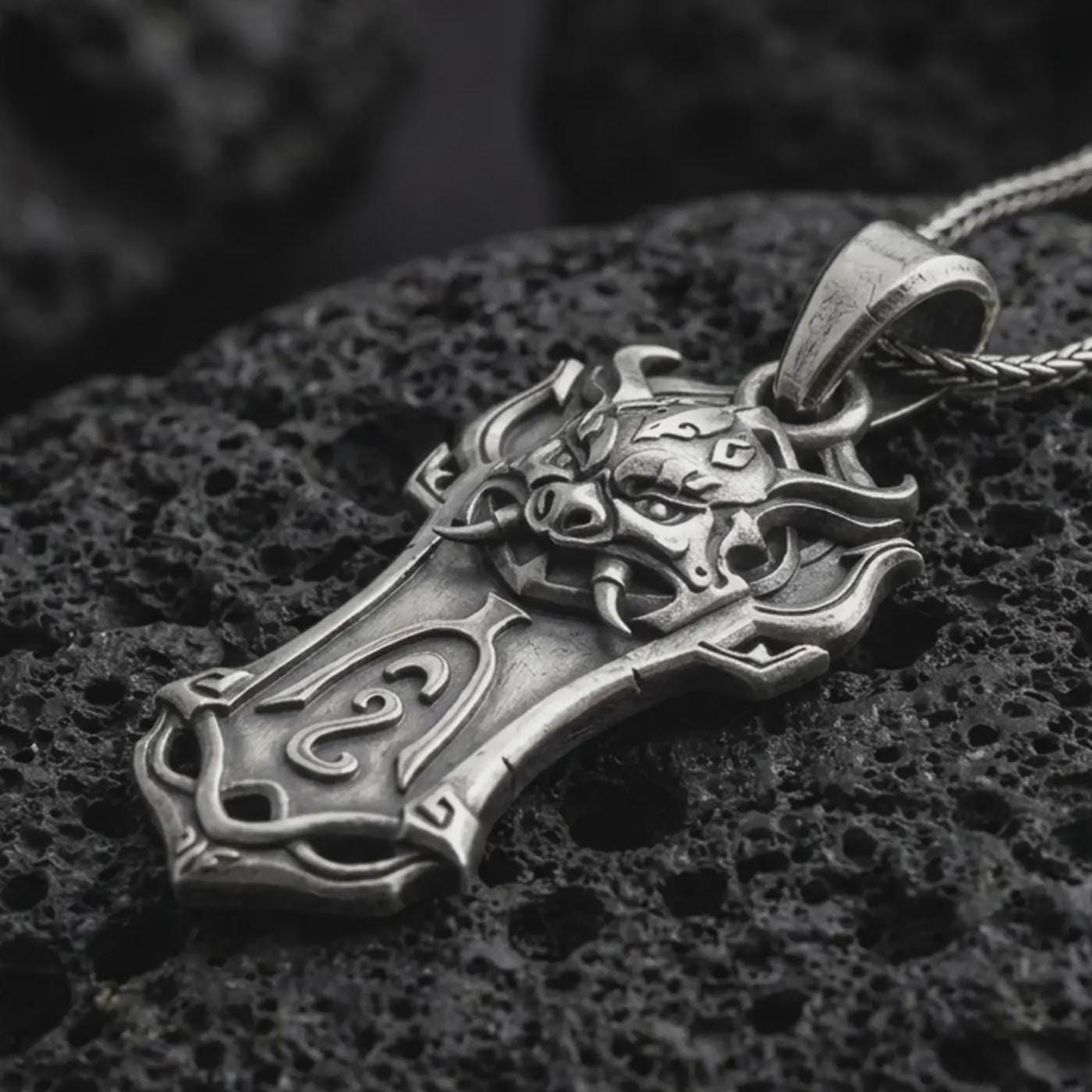 An oxidized Taurus bull pendant rests on dark volcanic rock in a macro studio close-up, showing a horned animal head over a shield-inspired frame.