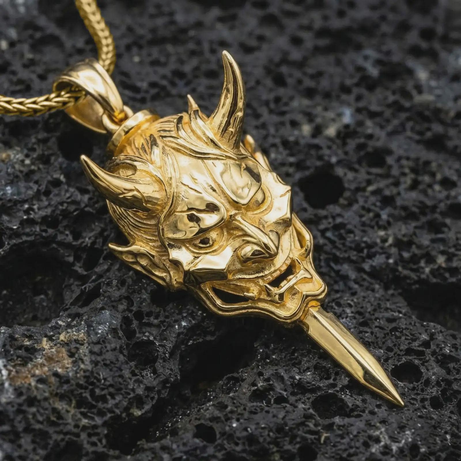 A gold plated oni mask pendant lies on textured volcanic rock in a dramatic studio macro shot, highlighting the protective demon mask design.
