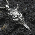 Load image into Gallery viewer, A polished silver oni mask pendant is displayed on black volcanic rock in a premium macro studio close-up with sharp Japanese warrior-inspired texture.
