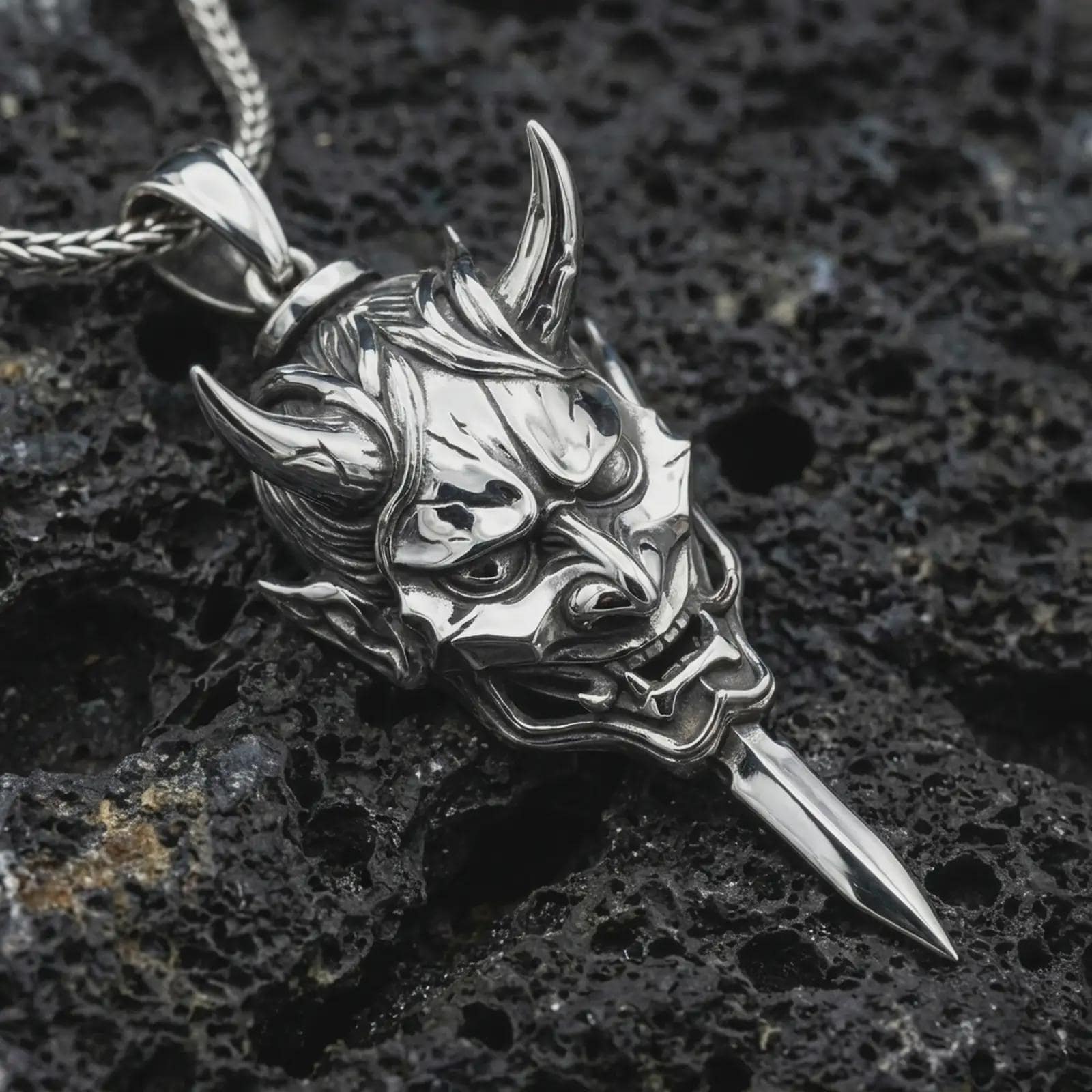 A polished silver oni mask pendant is displayed on black volcanic rock in a premium macro studio close-up with sharp Japanese warrior-inspired texture.