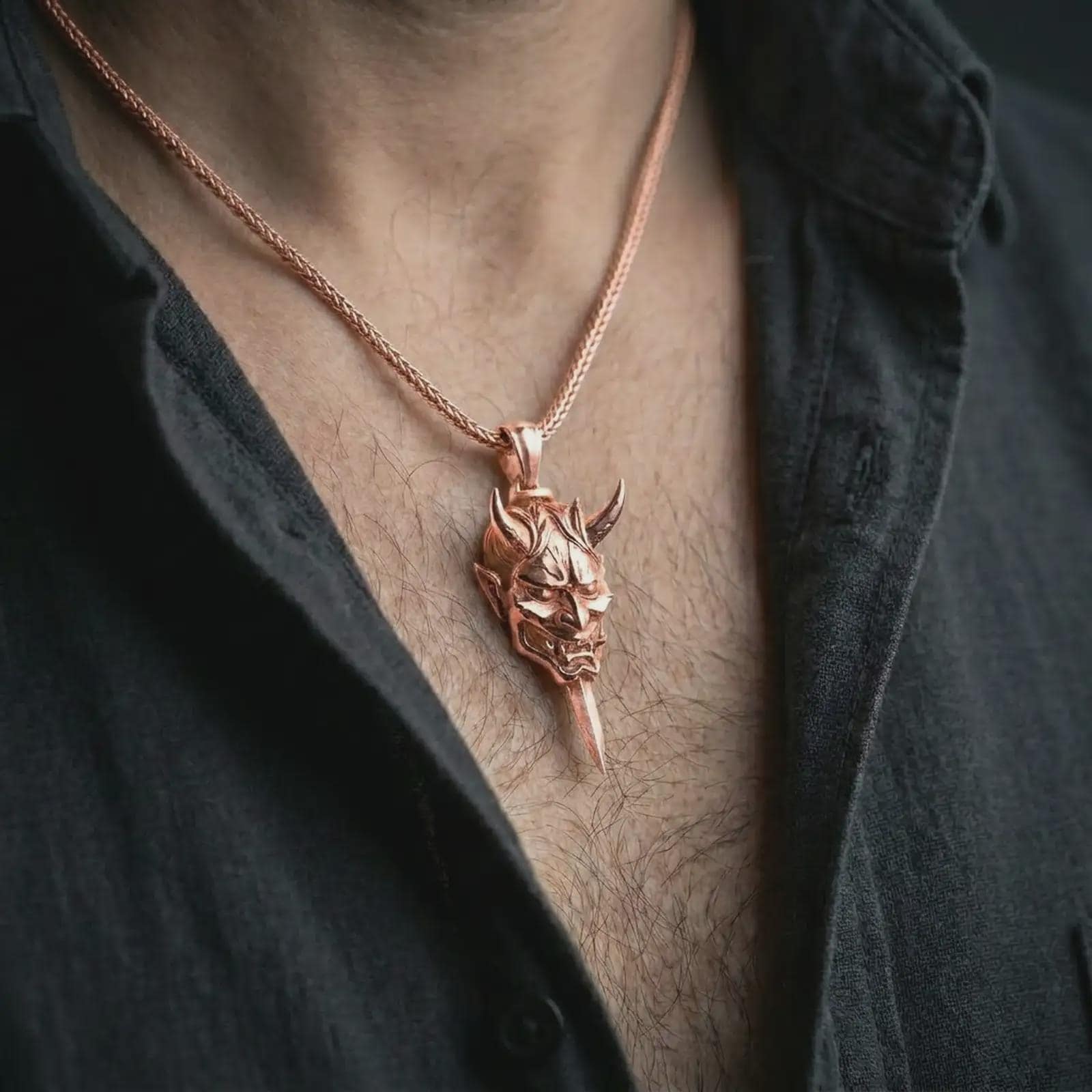 A rose gold plated oni mask pendant is worn on the neck in a close-up lifestyle composition, highlighting its oni mask detail and blade-shaped design.