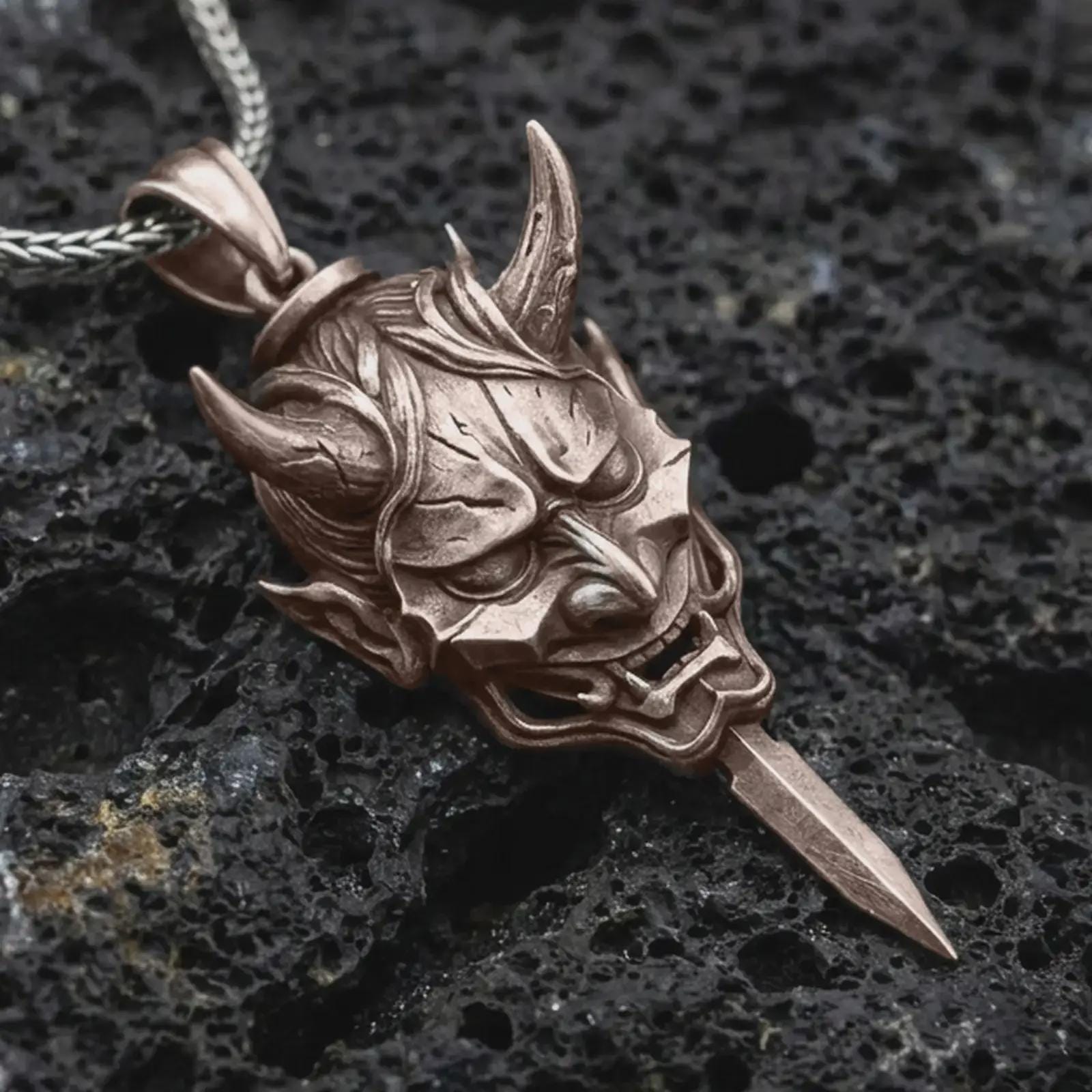A Bronze oni mask pendant rests on dark volcanic rock in a macro studio close-up, showing a fierce horned demon face with a blade-like detail.