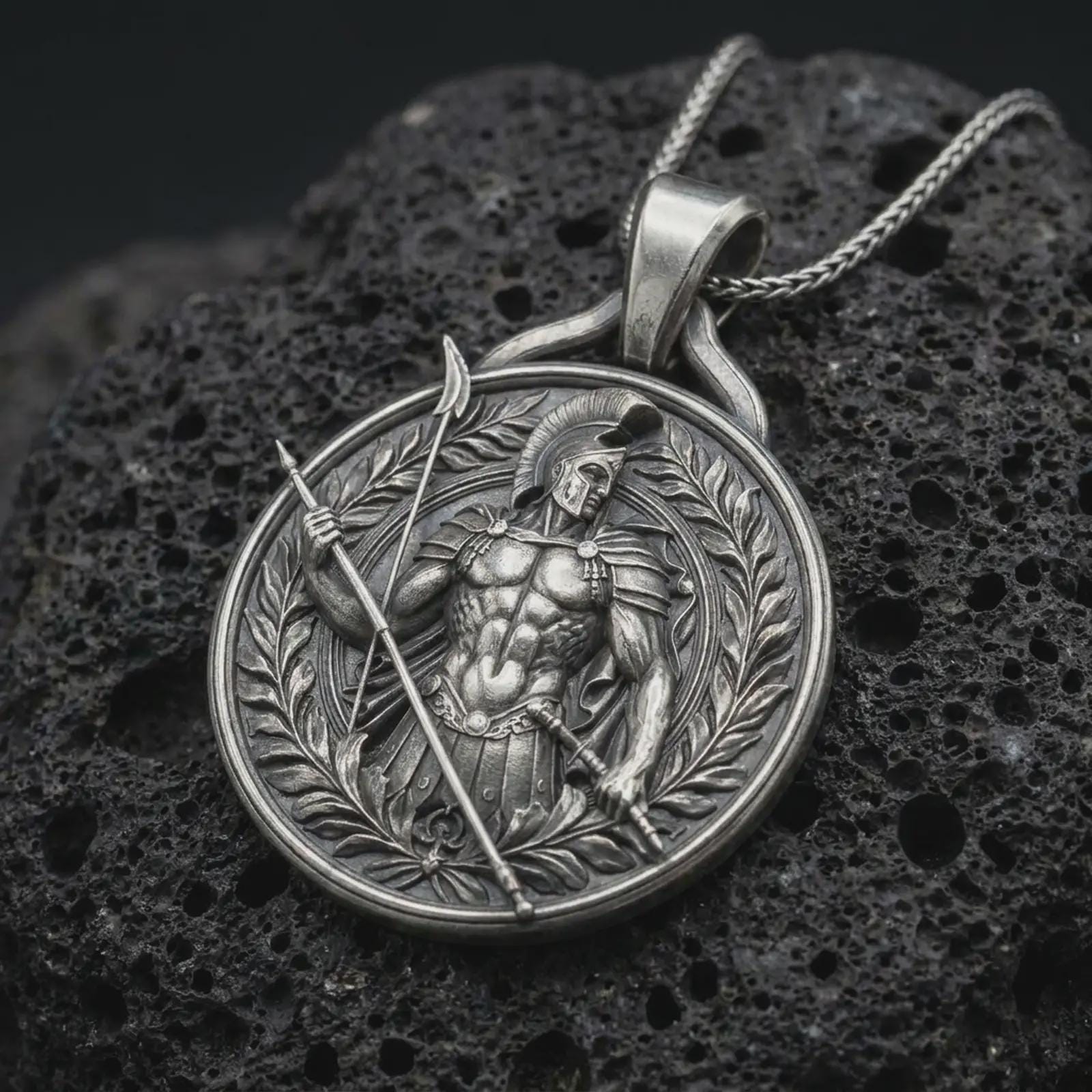An oxidized Spartan warrior pendant rests on dark volcanic rock in a macro studio close-up, showing a detailed Greek soldier with spear and laurel frame.