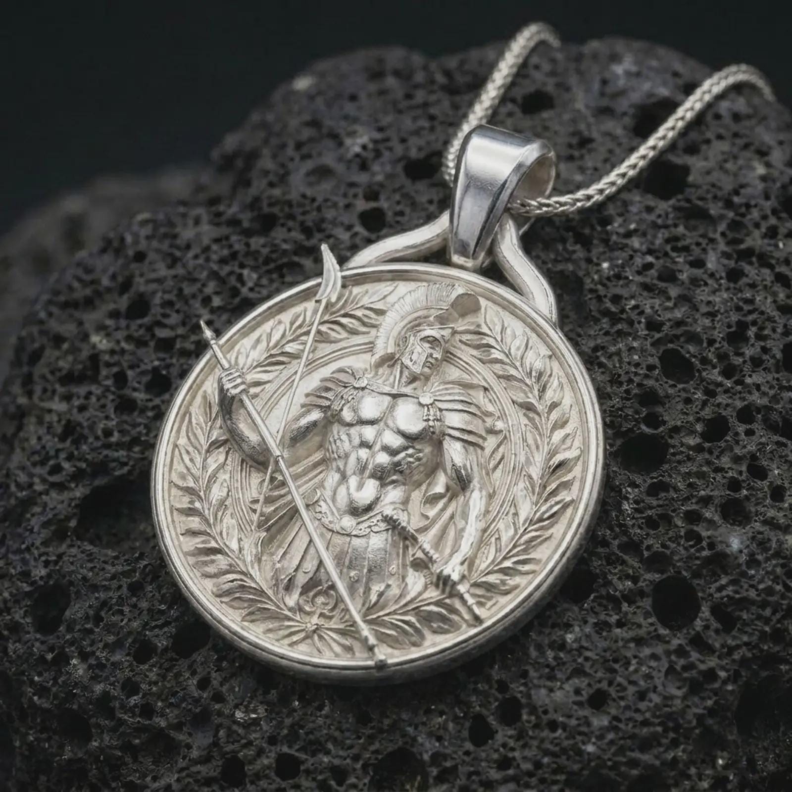 A polished silver Spartan warrior pendant is displayed on black volcanic rock in a premium macro studio close-up with sharp ancient soldier details.