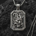 Load image into Gallery viewer, An oxidized Hannya mask pendant rests on dark volcanic rock in a macro studio close-up, showing a fierce Japanese demon face inside an ornate frame.
