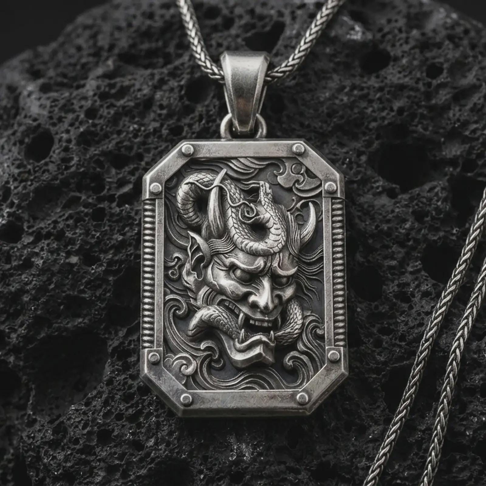 An oxidized Hannya mask pendant rests on dark volcanic rock in a macro studio close-up, showing a fierce Japanese demon face inside an ornate frame.