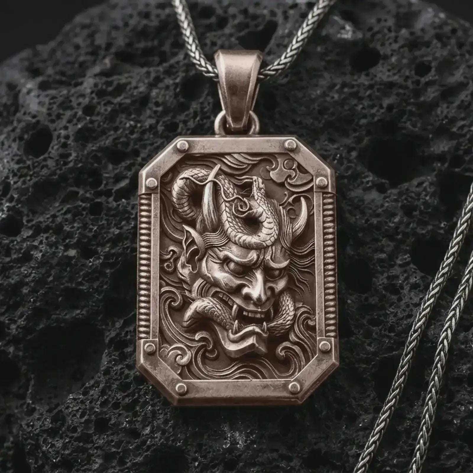 A Bronze Hannya mask pendant rests on dark volcanic rock in a macro studio close-up, showing a fierce Japanese demon face inside an ornate frame.