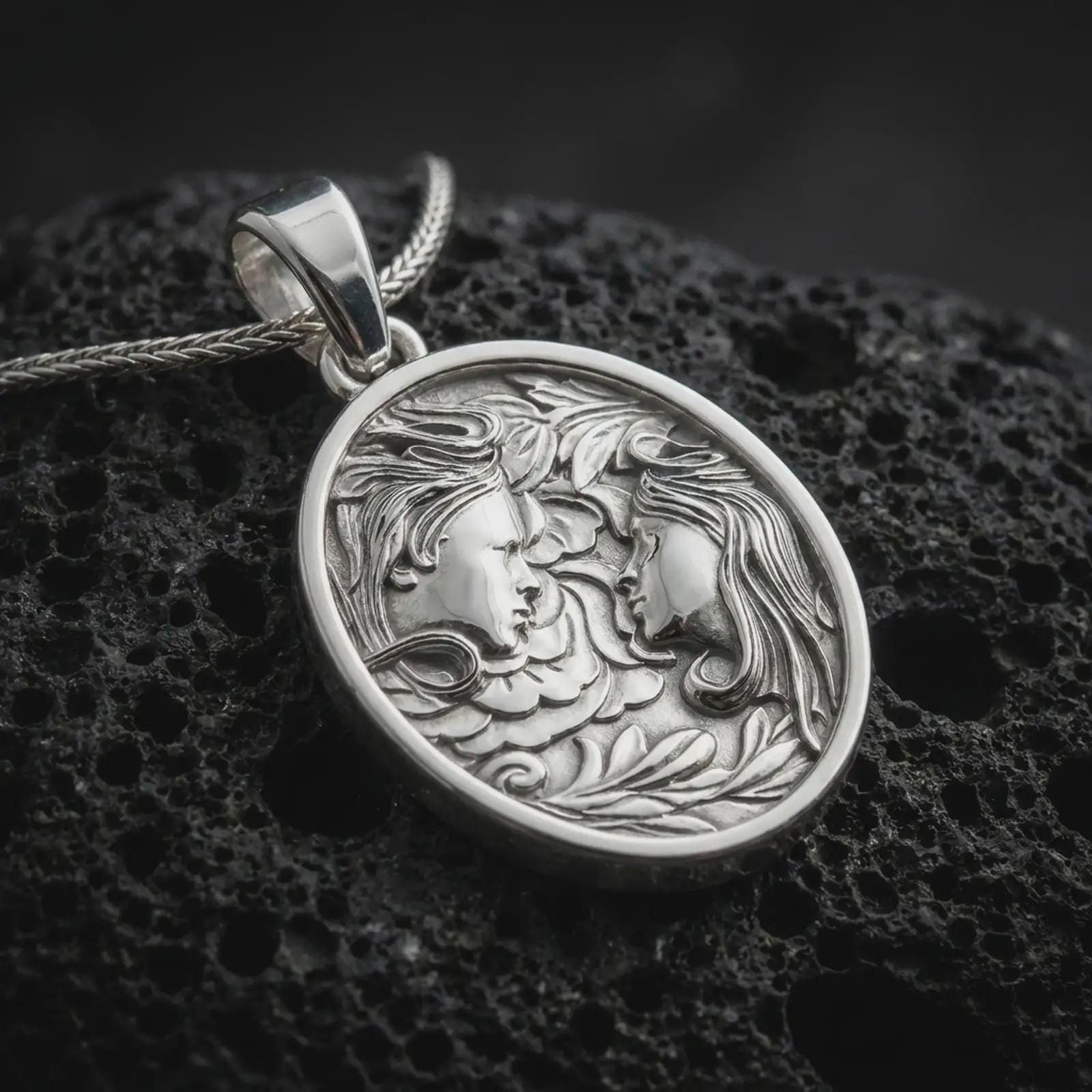 A polished silver Gemini pendant is displayed on black volcanic rock in a premium macro studio close-up with detailed twin faces and flowing botanical texture.
