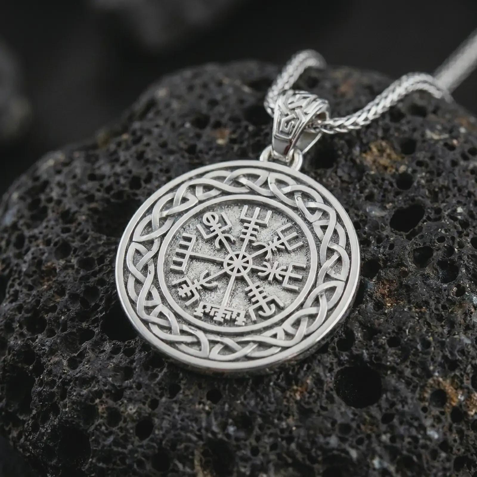 Polished silver Vegvisir Viking compass pendant necklace on textured volcanic rock, centered macro jewelry photo with detailed runic compass engraving and bright silver highlights.