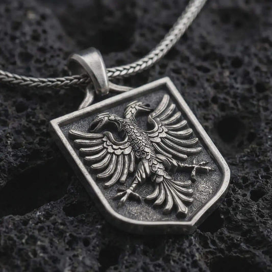 Oxidized double-headed eagle shield pendant necklace on textured volcanic rock, close-up studio shot highlighting the twin eagle heads, spread wings, crest frame, and antique silver contrast.