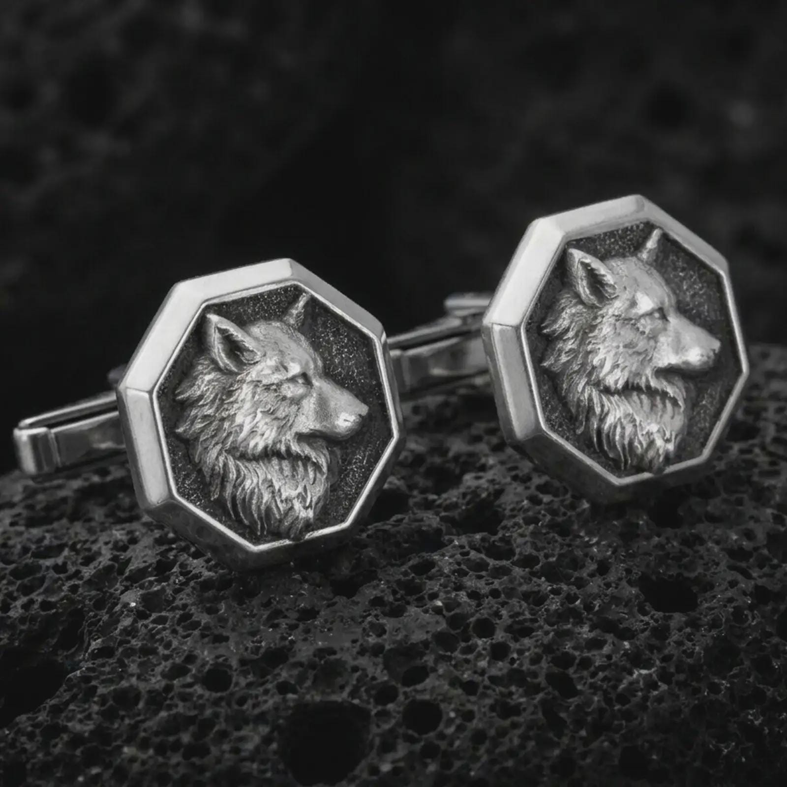 An oxidized wolf head cufflink pair rests on dark volcanic rock in a macro studio close-up, showing sculpted animal detail inside octagon frames.