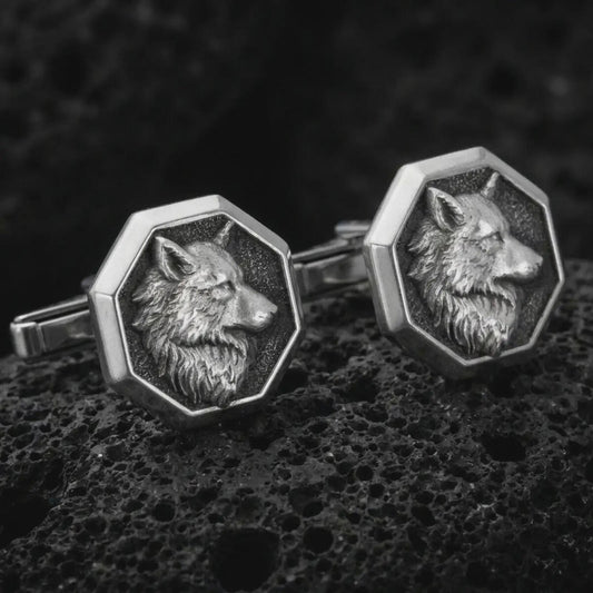 An oxidized wolf head cufflink pair rests on dark volcanic rock in a macro studio close-up, showing sculpted animal detail inside octagon frames.