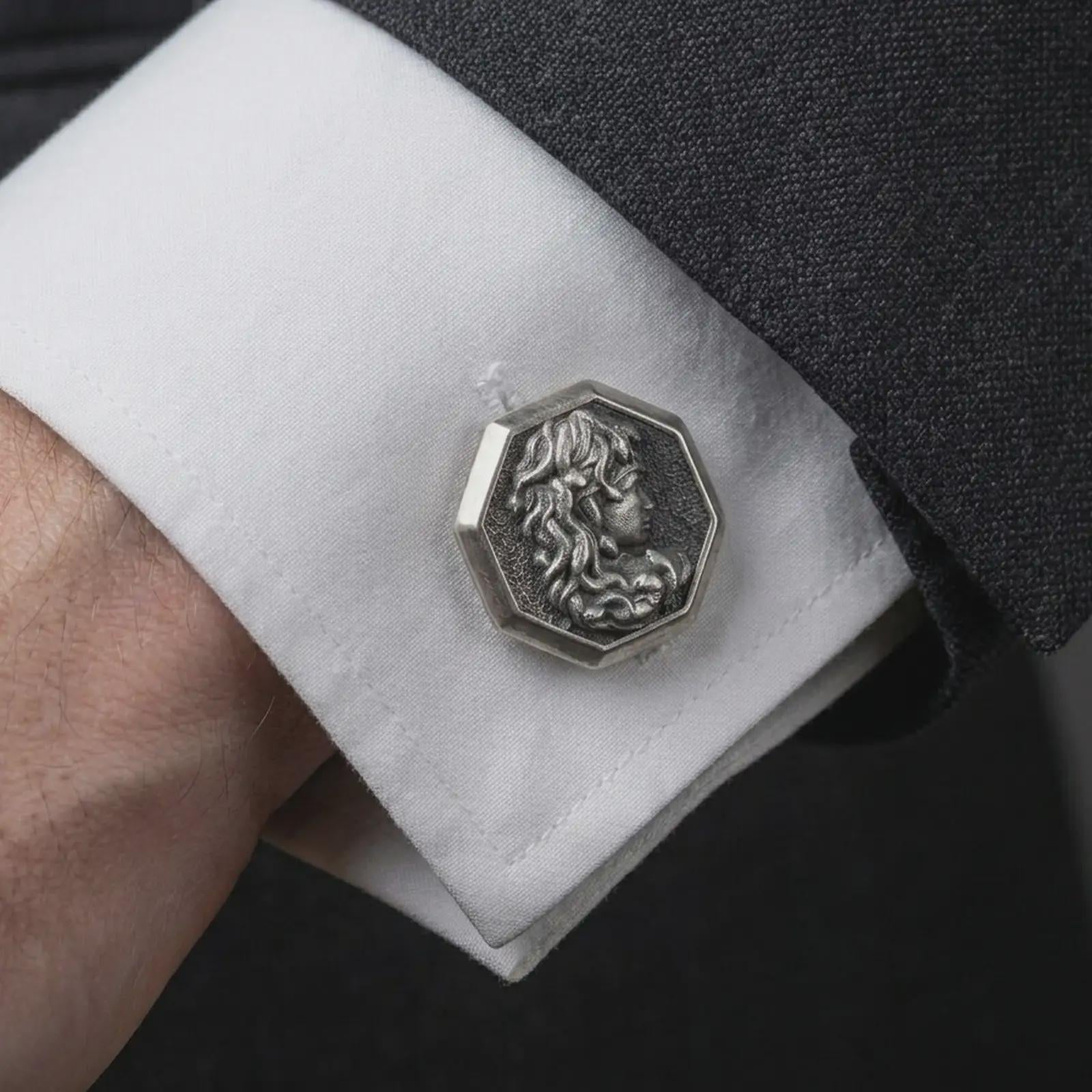 An oxidized Medusa cufflink is worn on a white shirt cuff in a close-up lifestyle shot, styled with a dark suit for a powerful formal look.