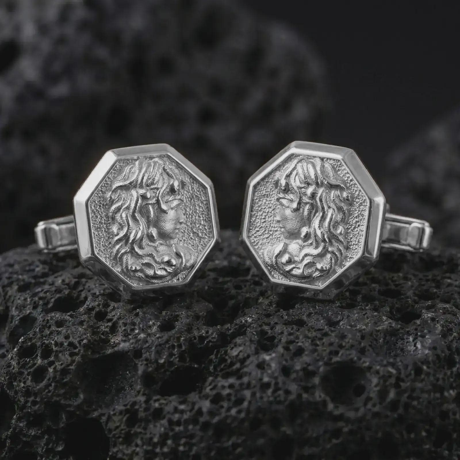 Polished silver Medusa cufflinks are displayed on black volcanic rock in a premium macro studio close-up with detailed Greek mythology texture.