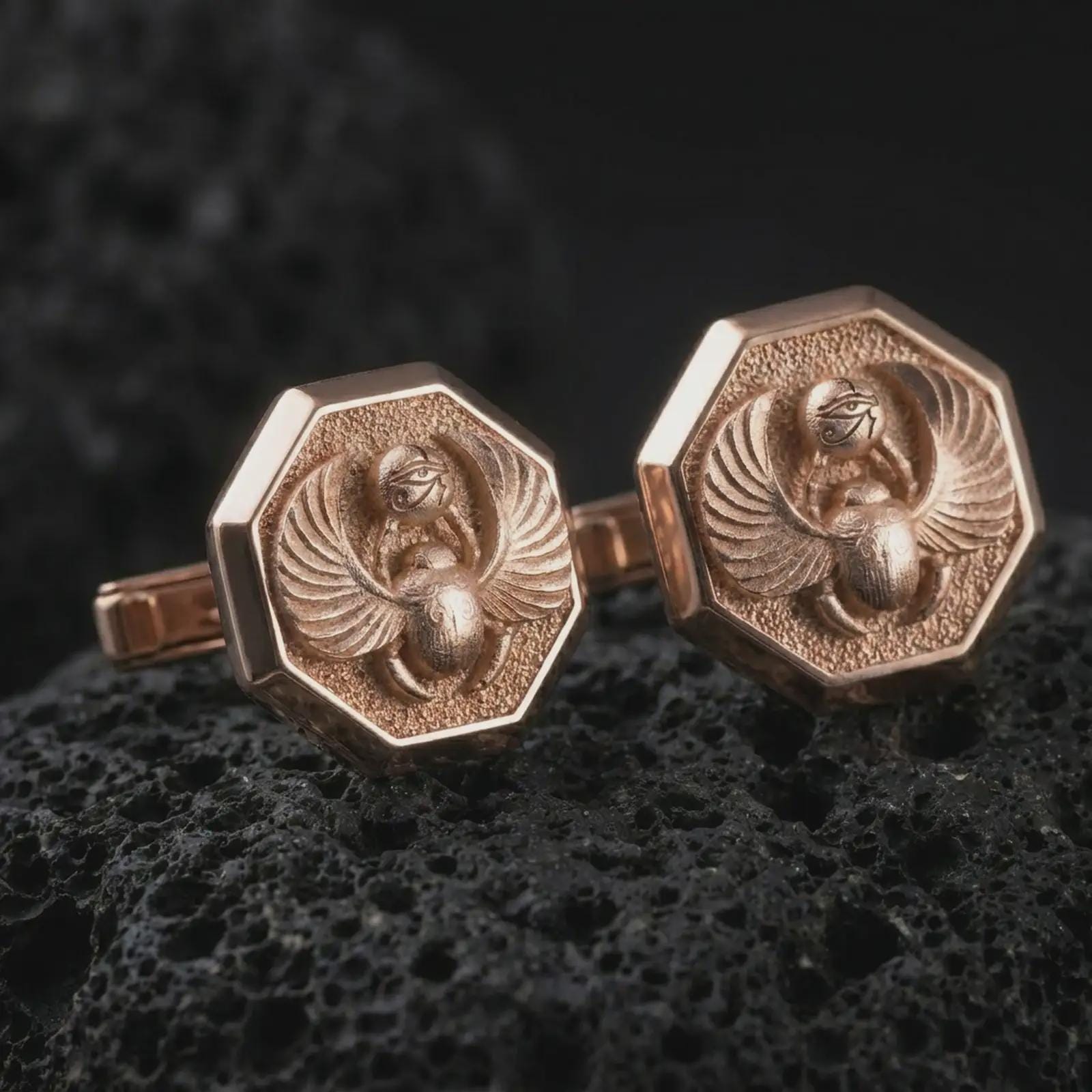 Rose gold plated scarab cufflinks are photographed on porous volcanic rock in a close-up studio composition with refined luxury lighting and bold ancient detail.