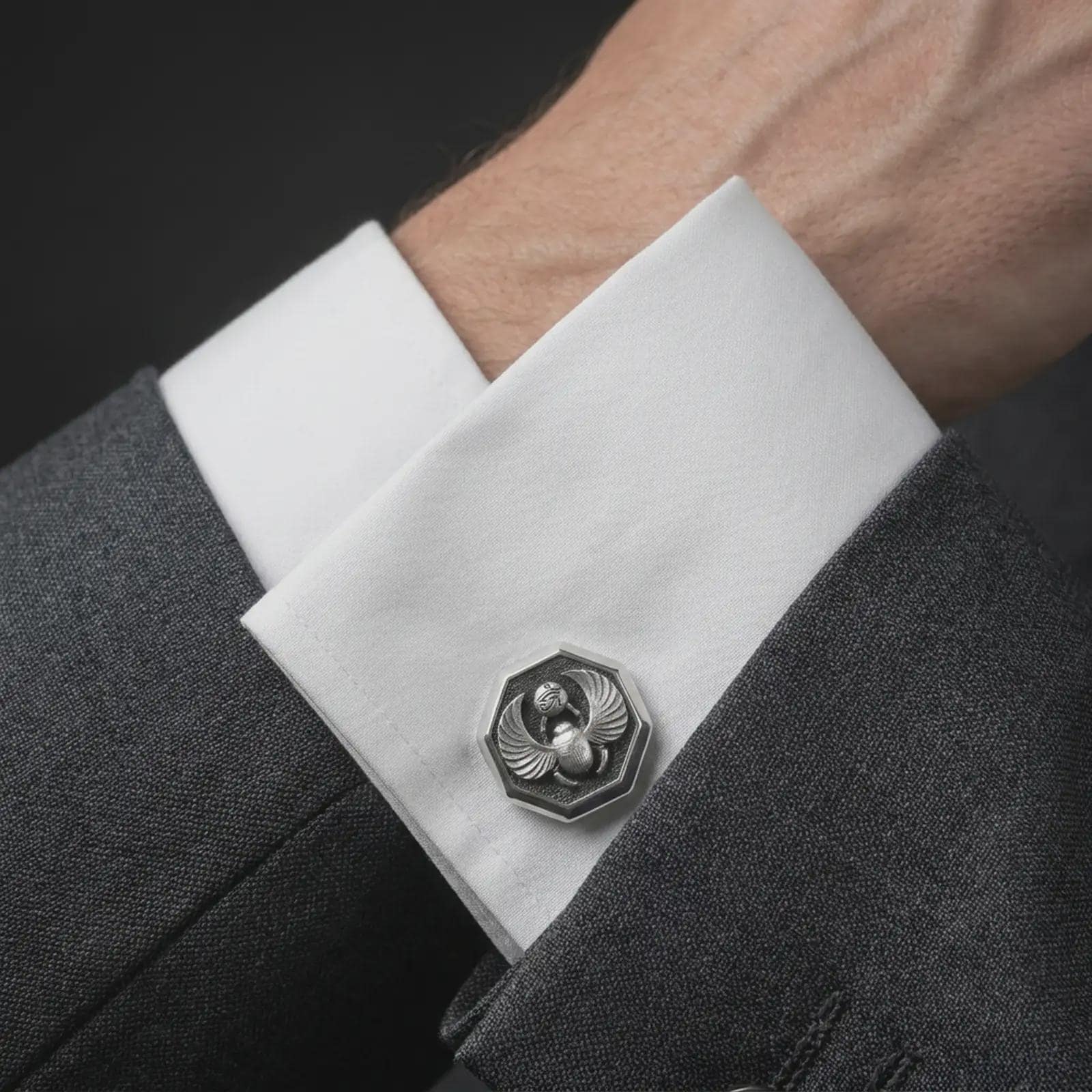 An oxidized scarab cufflink is worn on a white shirt cuff in a close-up lifestyle shot, styled with a dark suit as a symbolic Egyptian accessory.