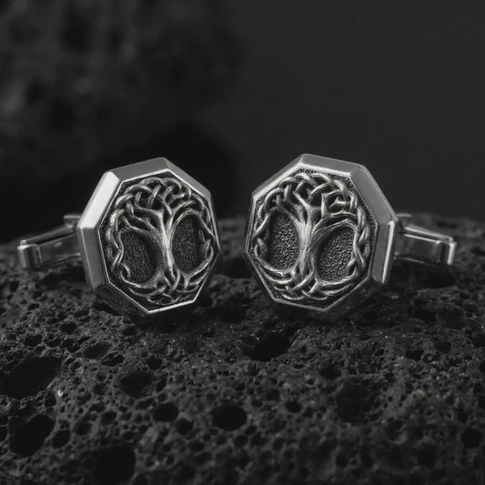An oxidized Yggdrasil cufflink pair rests on dark volcanic rock in a macro studio close-up, showing a woven Tree of Life design inside octagon frames.