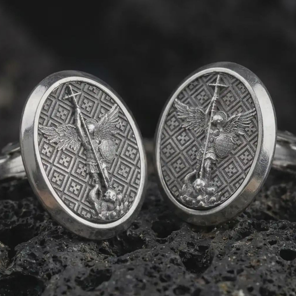 Oxidized Saint Michael cufflinks on textured volcanic rock, close-up studio shot highlighting the archangel relief, spear, wings, patterned background, and antique silver contrast.