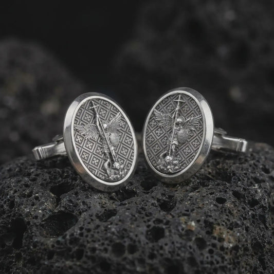 Oxidized Saint Michael cufflinks on textured volcanic rock, close-up studio shot highlighting the archangel relief, spear, wings, patterned background, and antique silver contrast.