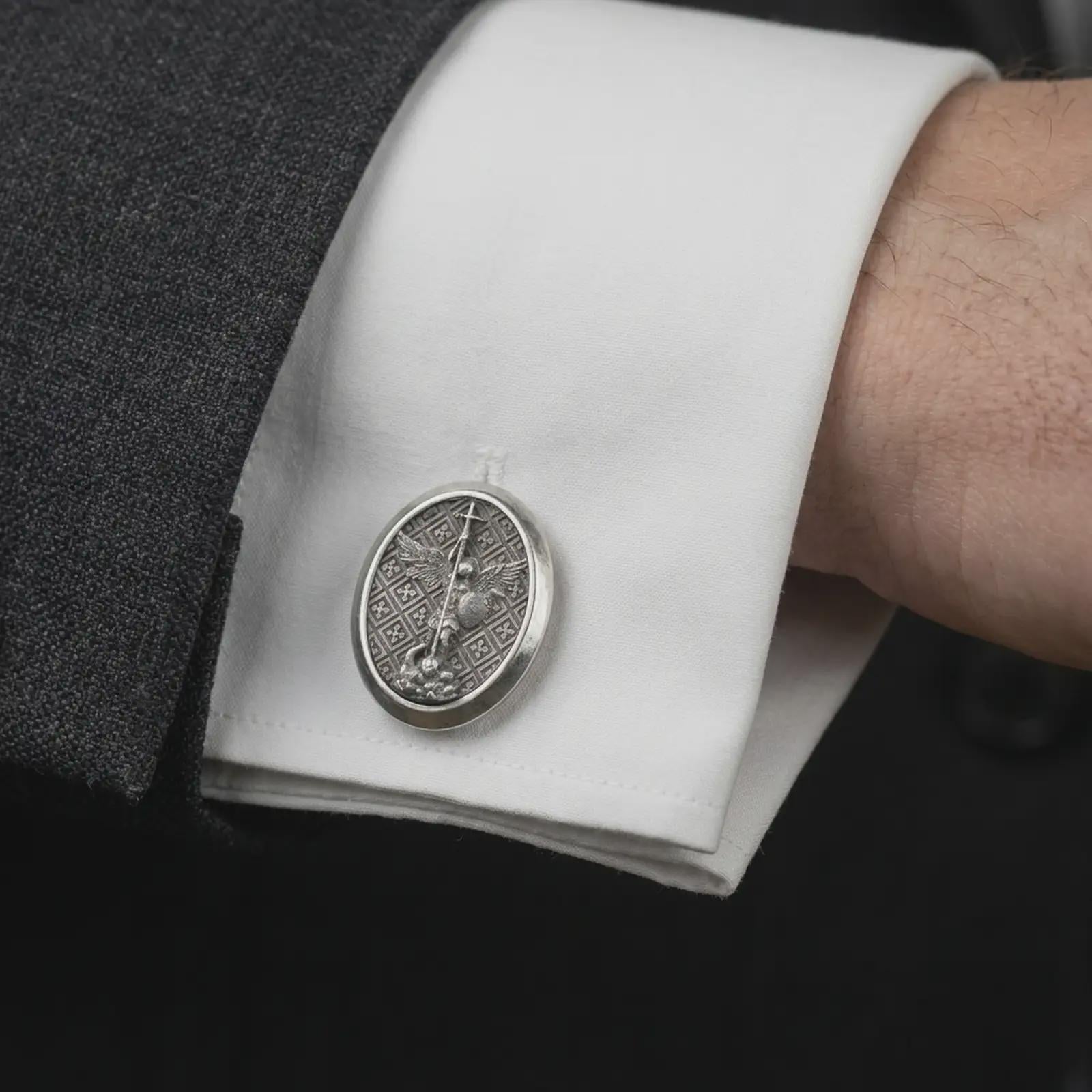 Oxidized Saint Michael cufflinks worn on a suit cuff, close-up lifestyle jewelry photo showing the guardian angel medal design with crisp white shirt fabric.