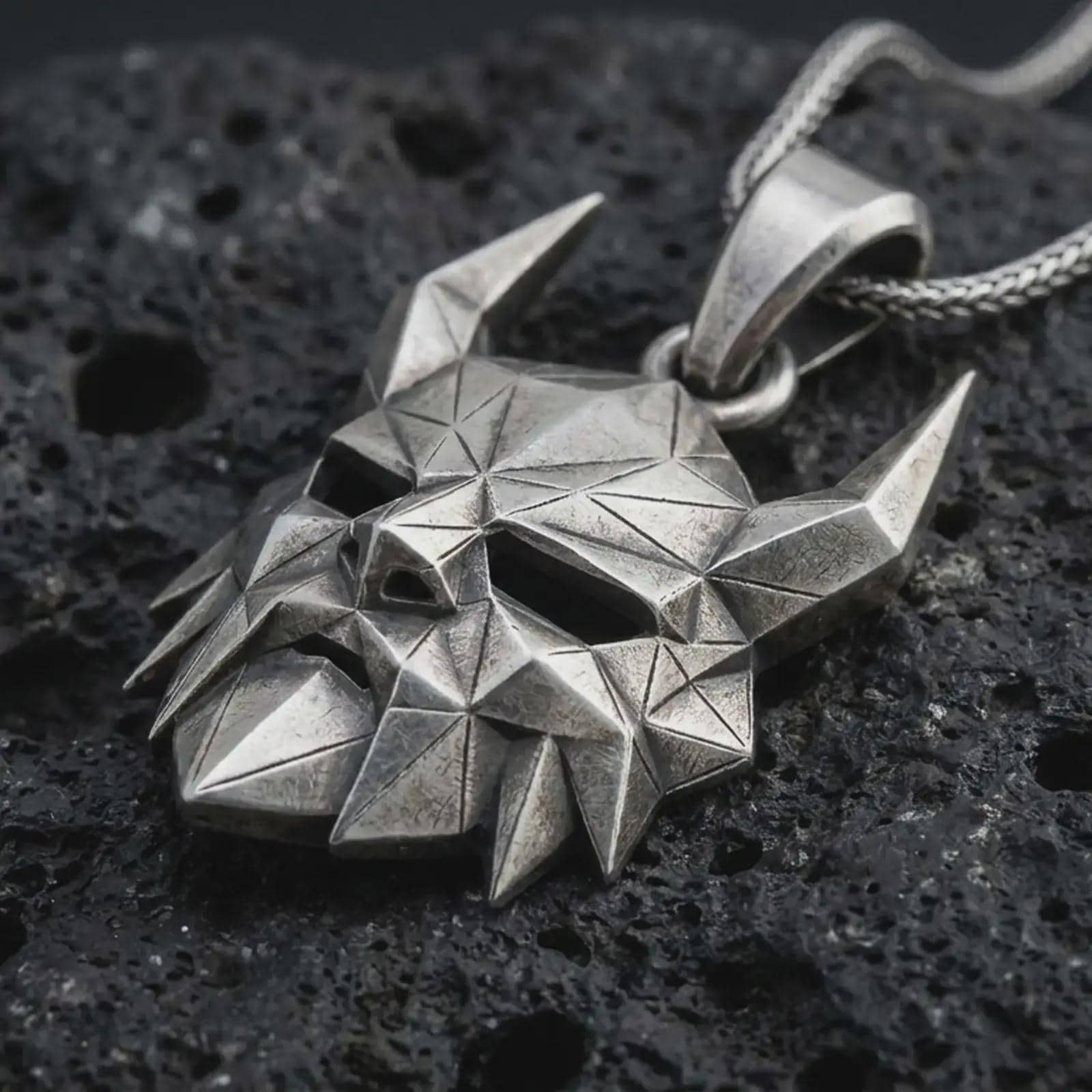 An oxidized Surtur pendant rests on dark volcanic rock in a macro studio close-up, showing a sharp horned mask with angular Norse fantasy detail.