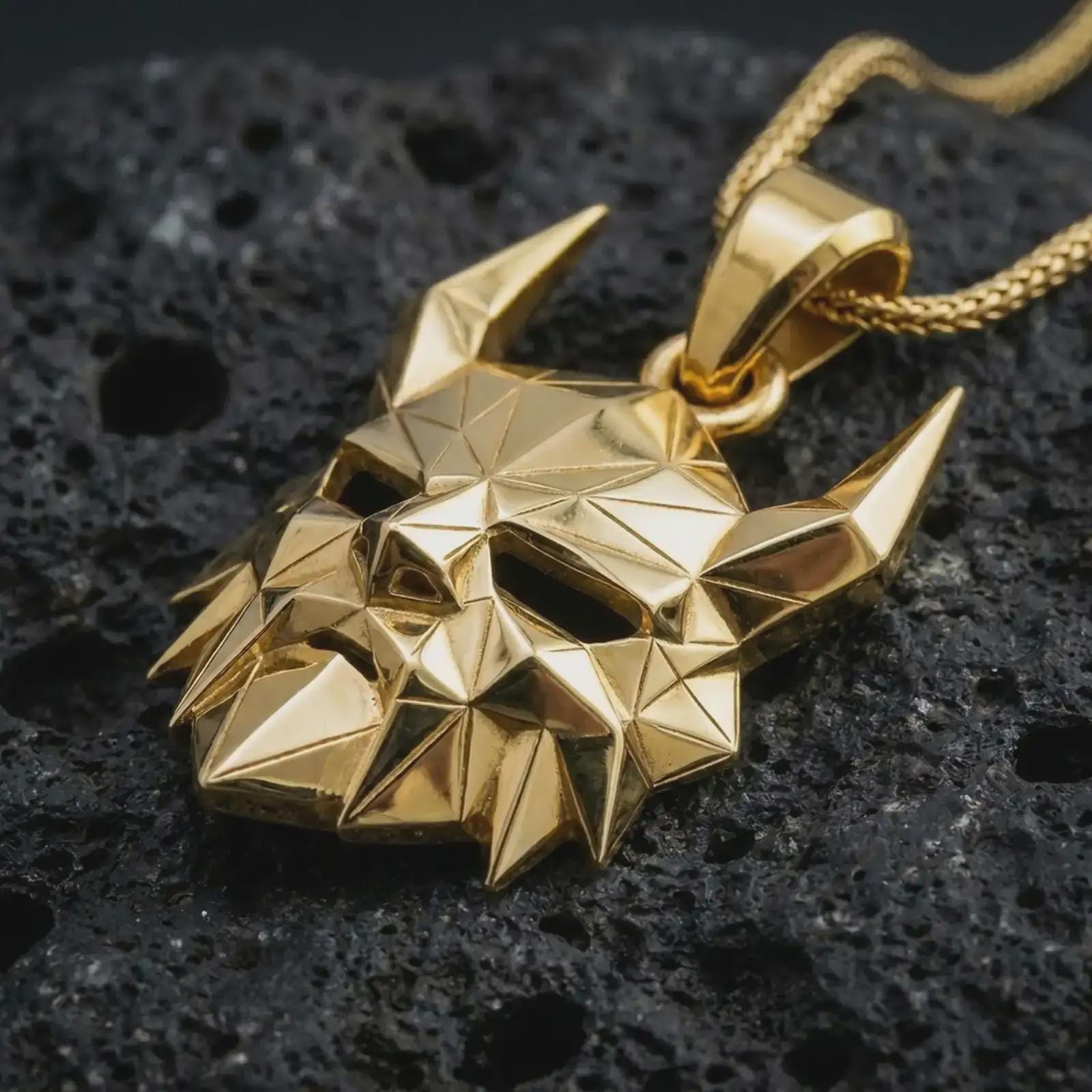 A gold plated Surtur pendant lies on textured volcanic rock in a dramatic studio macro shot, highlighting the horned mask motif and Ragnarok-inspired style.