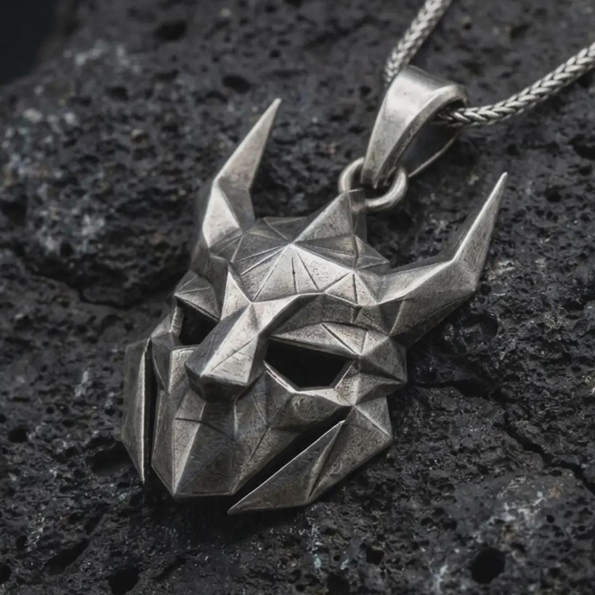An oxidized Loki pendant rests on dark volcanic rock in a macro studio close-up, showing a faceted horned mask inspired by Norse mythology.