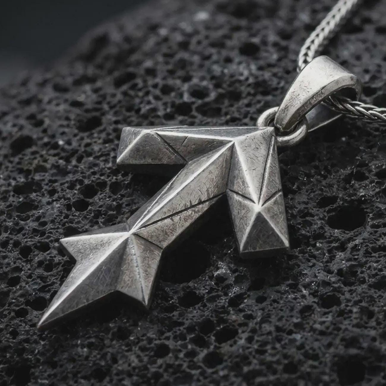 An oxidized Tyr rune pendant rests on dark volcanic rock in a macro studio close-up, showing the Teiwaz symbol in a bold geometric Norse design.