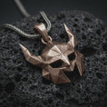 Load image into Gallery viewer, A bronze Helm of Awe inspired mask pendant rests on dark volcanic rock in a macro studio close-up, showing a faceted horned Norse warrior design.
