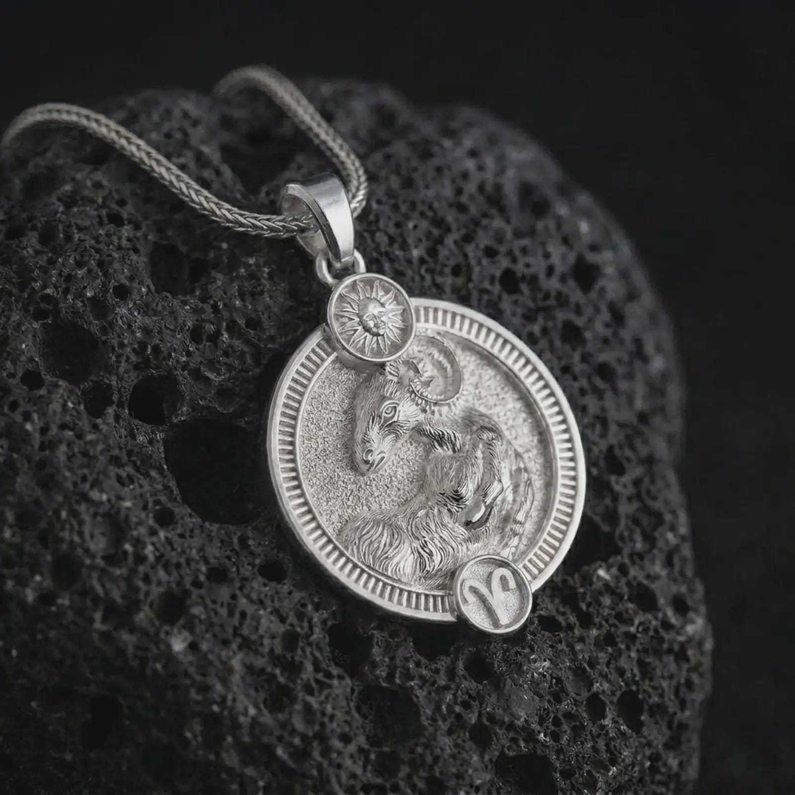 Polished silver Aries zodiac pendant necklace on textured volcanic rock, centered macro jewelry photo with detailed ram horoscope relief and bright silver highlights.