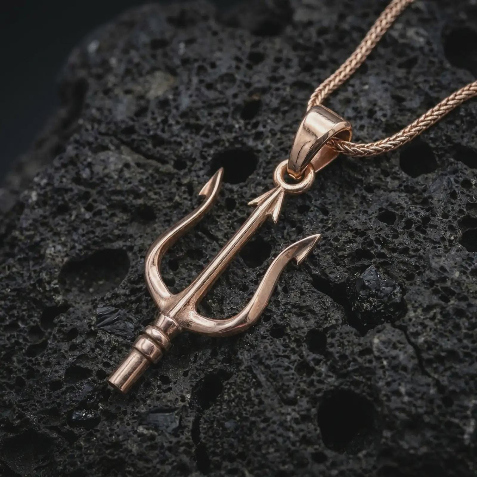 Rose gold plated Poseidon trident pendant necklace on textured volcanic rock, detailed macro product shot with soft warm reflections and ancient sea god symbolism.