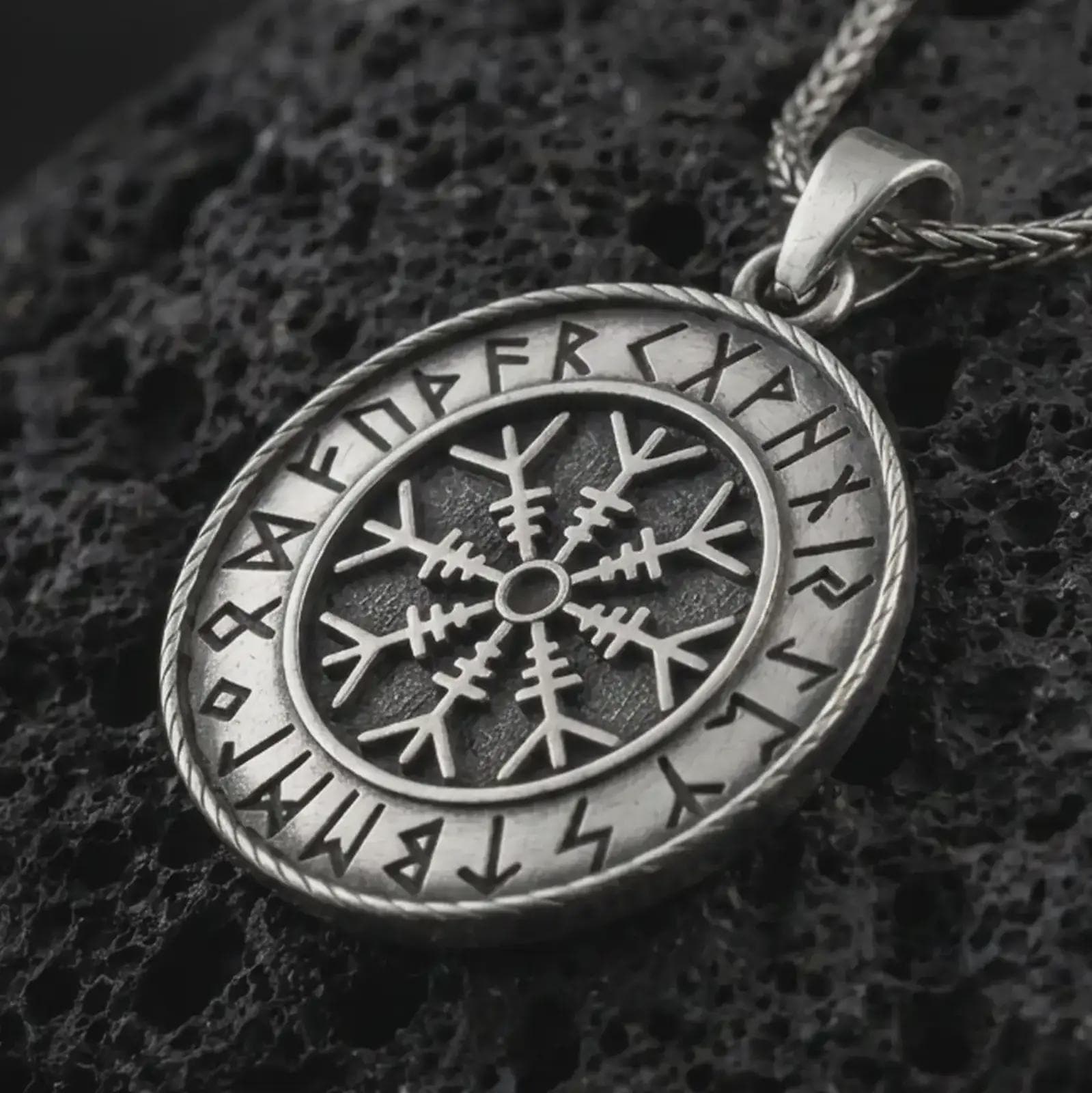 Oxidized Vegvisir Viking rune pendant necklace on textured volcanic rock, close-up studio shot highlighting the Norse compass symbol, rune border, rope frame, and antique silver contrast.