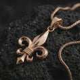 Load image into Gallery viewer, Rose gold plated fleur-de-lis pendant necklace on textured volcanic rock, detailed macro product shot with soft warm reflections and symbolic French lily styling.
