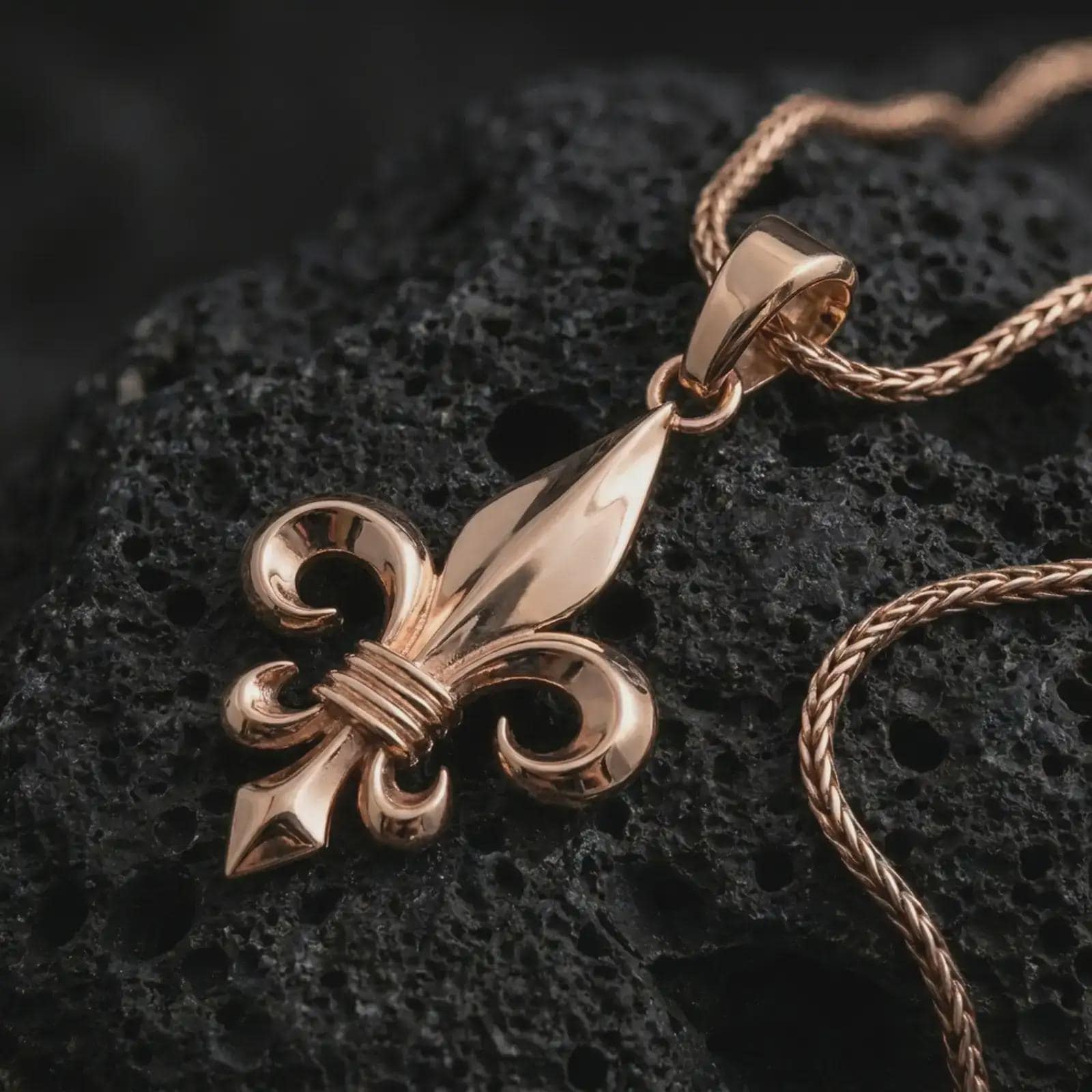 Rose gold plated fleur-de-lis pendant necklace on textured volcanic rock, detailed macro product shot with soft warm reflections and symbolic French lily styling.
