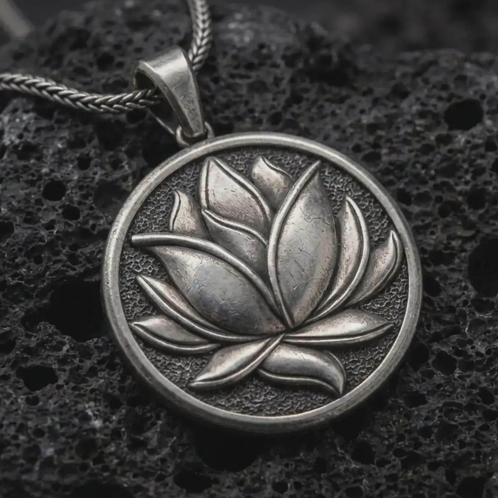Oxidized lotus flower pendant necklace on textured volcanic rock, close-up studio shot highlighting the layered petals, round medallion frame, textured background, and antique silver contrast.