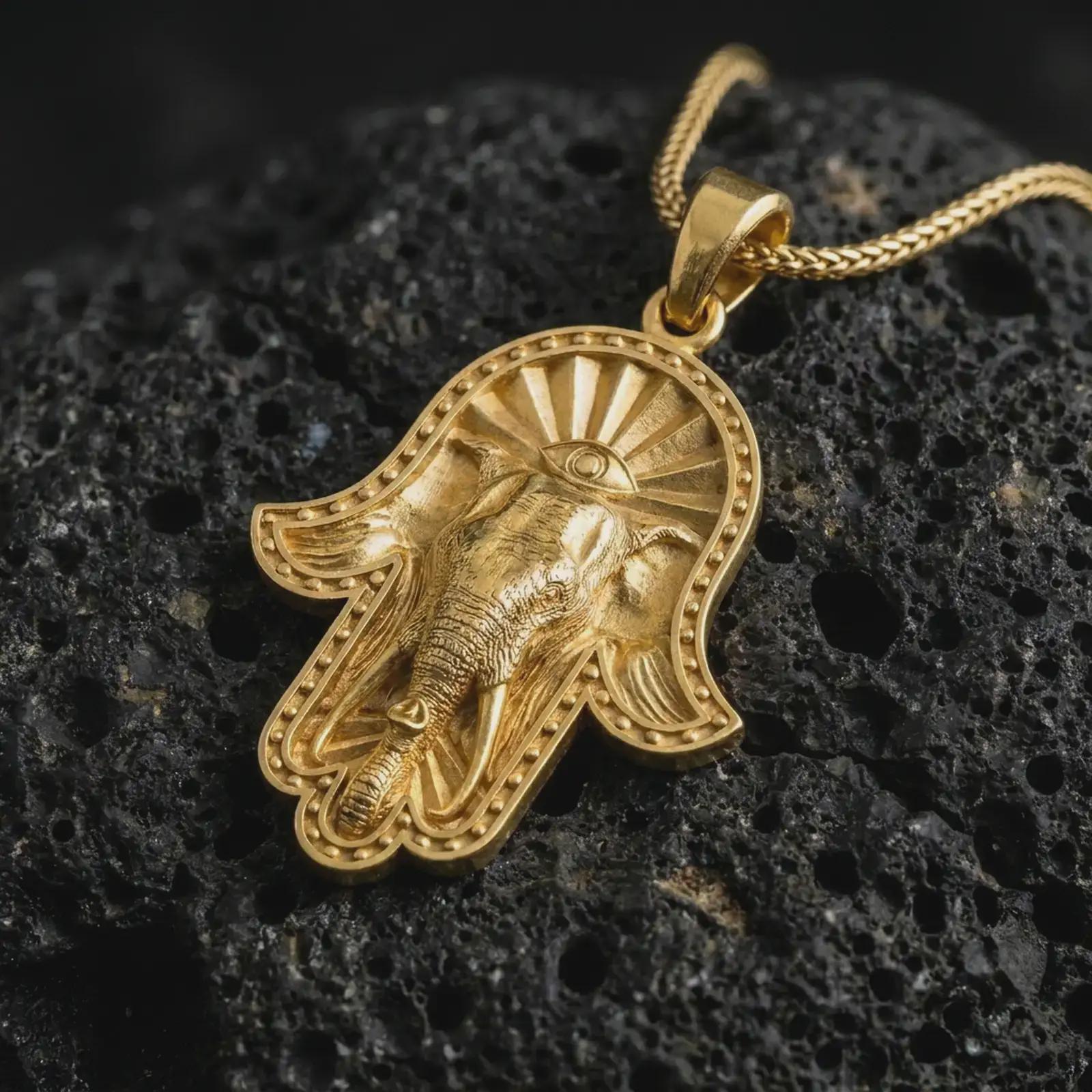 Gold plated hamsa elephant pendant necklace on textured volcanic rock, premium close-up studio composition emphasizing the protective hand shape, elephant symbolism, and sacred third eye design.