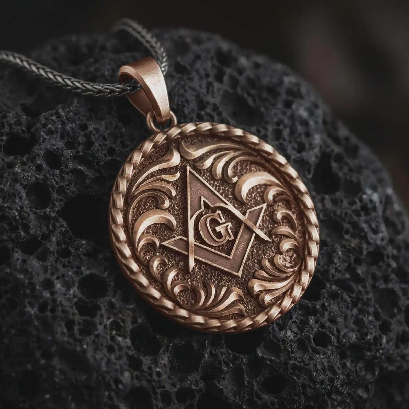 Bronze Masonic pendant necklace on textured volcanic rock, close-up studio shot highlighting the square and compass symbol, central G letter, ornate scrollwork, and antique silver contrast.