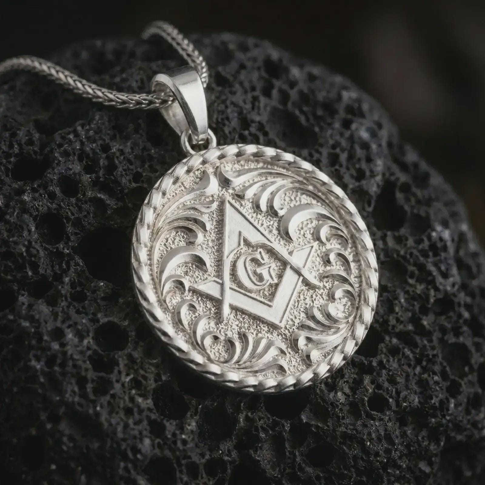 Polished silver Masonic pendant necklace on textured volcanic rock, centered macro jewelry photo with detailed Freemason emblem, textured background, and bright silver highlights.