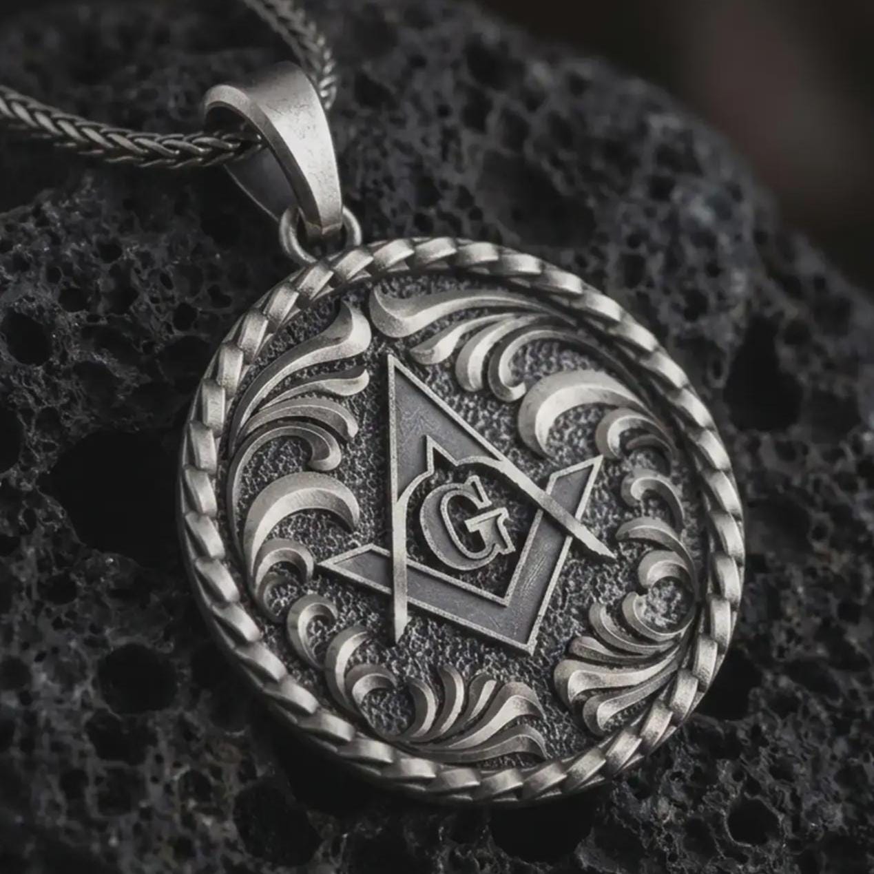 Oxidized Masonic pendant necklace on textured volcanic rock, close-up studio shot highlighting the square and compass symbol, central G letter, ornate scrollwork, and antique silver contrast.