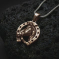 Load image into Gallery viewer, Bronze horseshoe pendant necklace on textured volcanic rock, close-up studio shot highlighting the horse head relief, lucky horseshoe frame, openwork design, and antique silver contrast.
