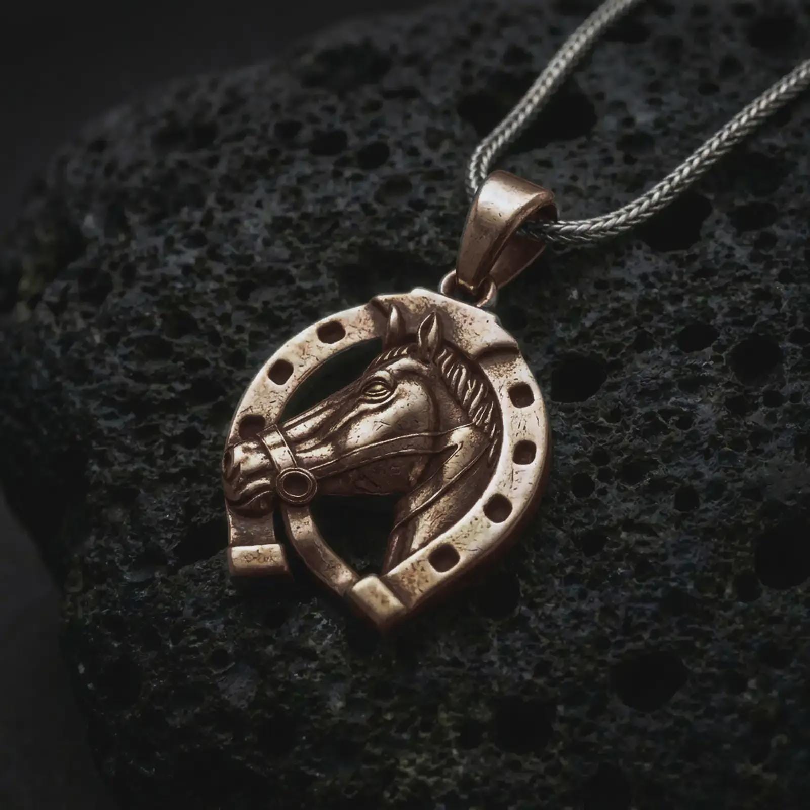 Bronze horseshoe pendant necklace on textured volcanic rock, close-up studio shot highlighting the horse head relief, lucky horseshoe frame, openwork design, and antique silver contrast.
