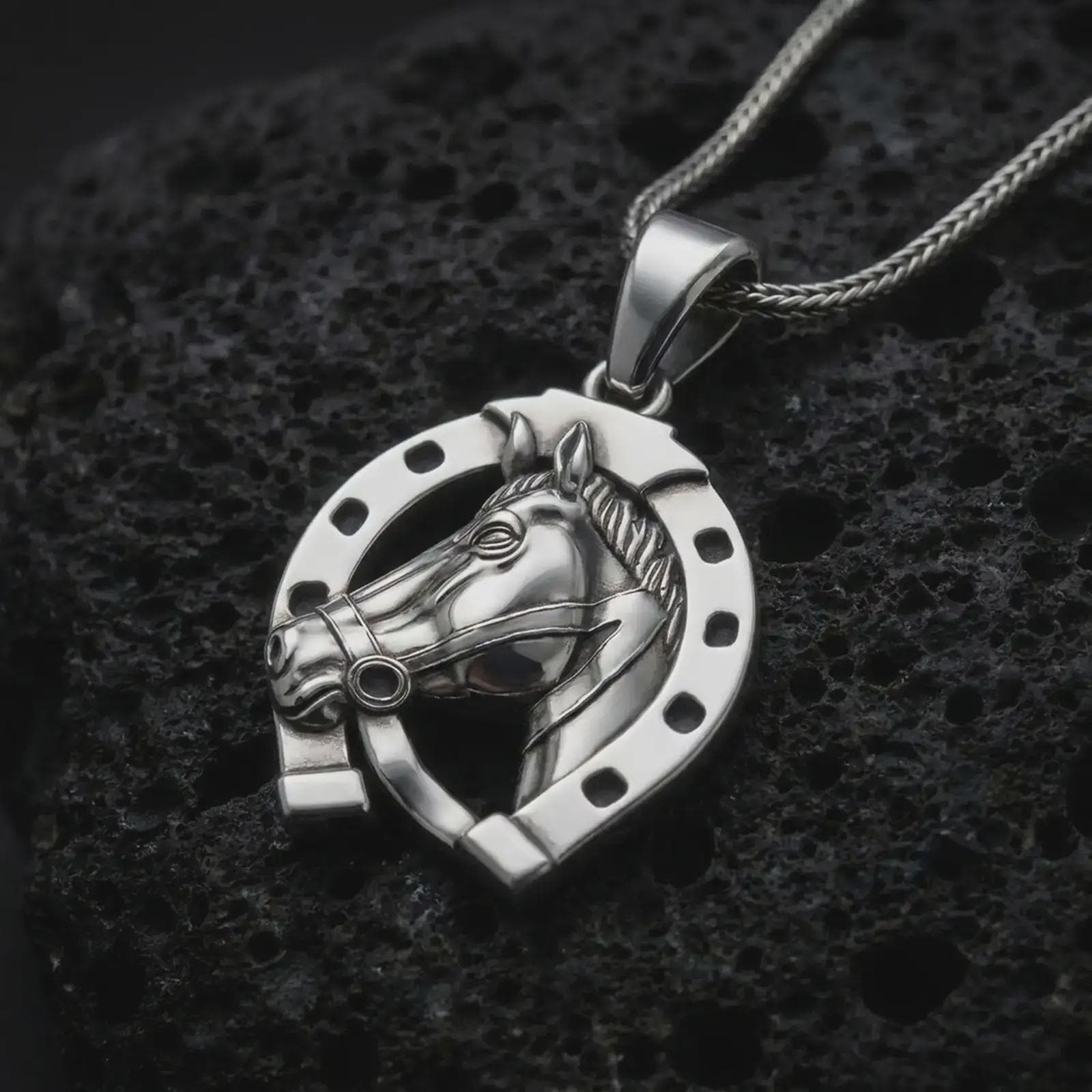 Polished silver horseshoe pendant necklace on textured volcanic rock, centered macro jewelry photo with detailed horse profile, bridle accents, and bright silver highlights.