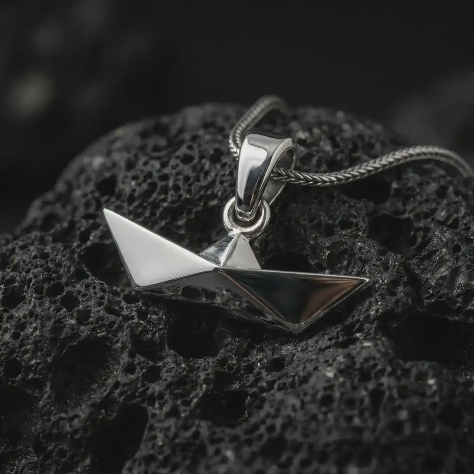 Polished silver origami boat pendant necklace on textured volcanic rock, centered macro jewelry photo with clean folded lines, angular silhouette, and bright silver highlights.