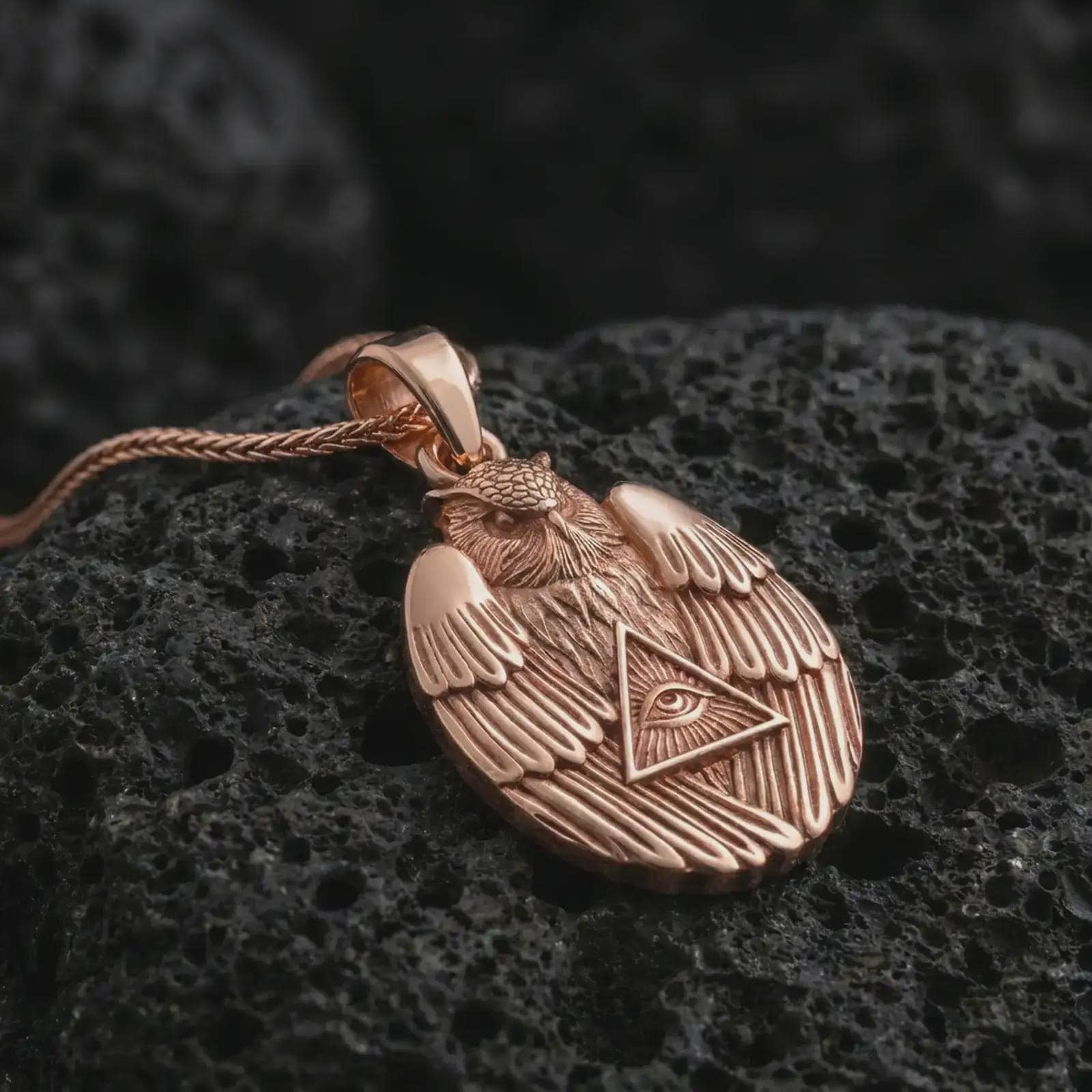 Rose gold plated owl all-seeing eye pendant necklace on textured volcanic rock, detailed macro product shot with soft warm reflections and symbolic owl protection styling.