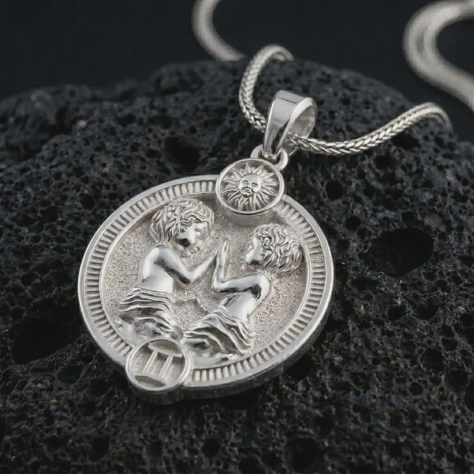 Polished silver Gemini zodiac pendant necklace on textured volcanic rock, centered macro jewelry photo with detailed horoscope twins relief and bright silver highlights.