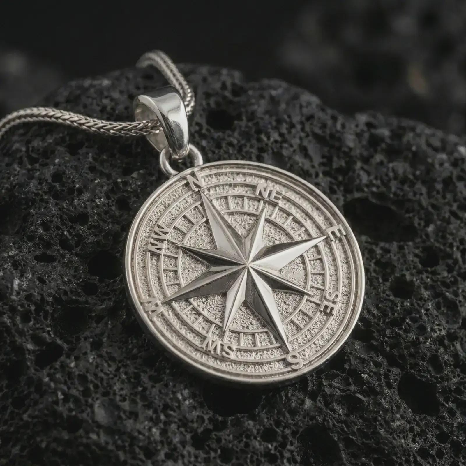 Polished silver compass rose pendant necklace on textured volcanic rock, centered macro jewelry photo with detailed navigation markings and bright silver highlights.