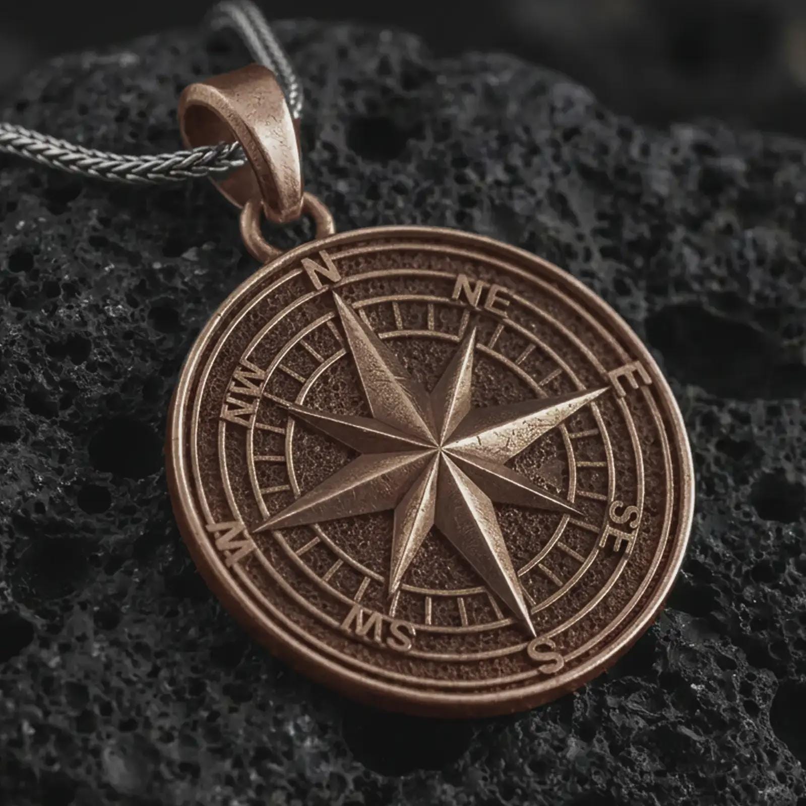 Bronze compass rose pendant necklace on textured volcanic rock, close-up studio shot highlighting the raised directional star, cardinal letters, circular map dial, and antique silver contrast.